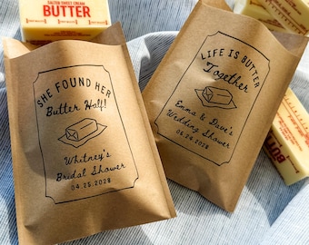 May include: Two brown paper bags featuring black printed text and illustrations. One bag displays She Found Her Butter Half! Whitney's Bridal Shower 04.25.2028. The other shows Life Is Butter Together Emma & Dave's Wedding Shower 06.24.2028. Several sticks of butter are present.
