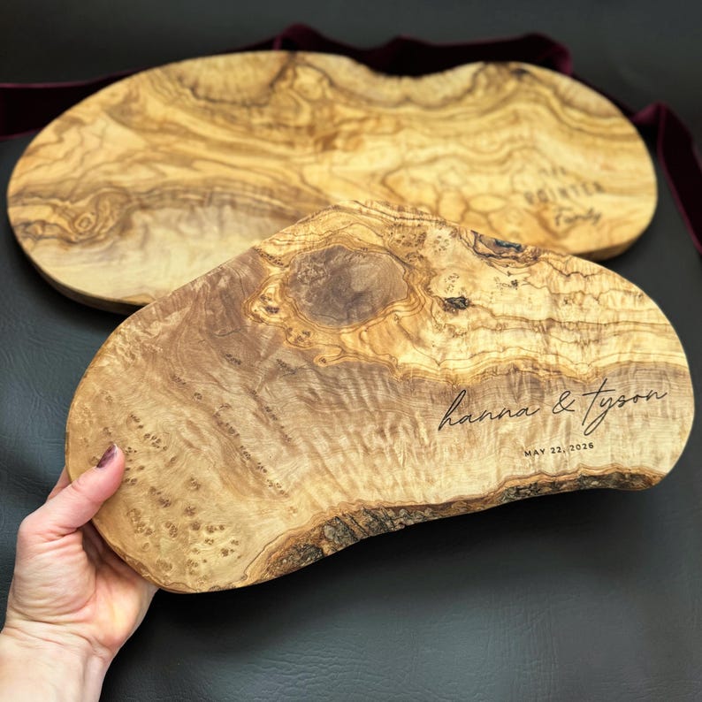 May include: Two olive wood serving boards with natural wood grain patterns. The front board is engraved with "hanna & tyson" and "MAY 22, 2026". The boards have a dark brown edge and are on a dark surface.