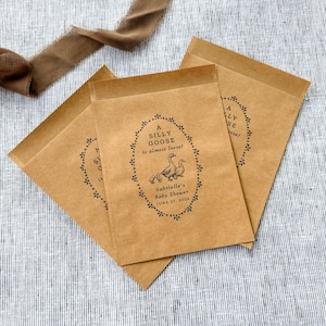 Puede incluir: Tres bolsas de papel marr&oacute;n para regalos con el texto "A SILLY GOOSE is almost loose!" en un marco ovalado decorativo. El marco incluye ilustraciones de gansos y gansitos. El texto "Gabrielle's Baby Shower JUNE 27, 2026" tambi&eacute;n est&aacute; impreso en las bolsas.