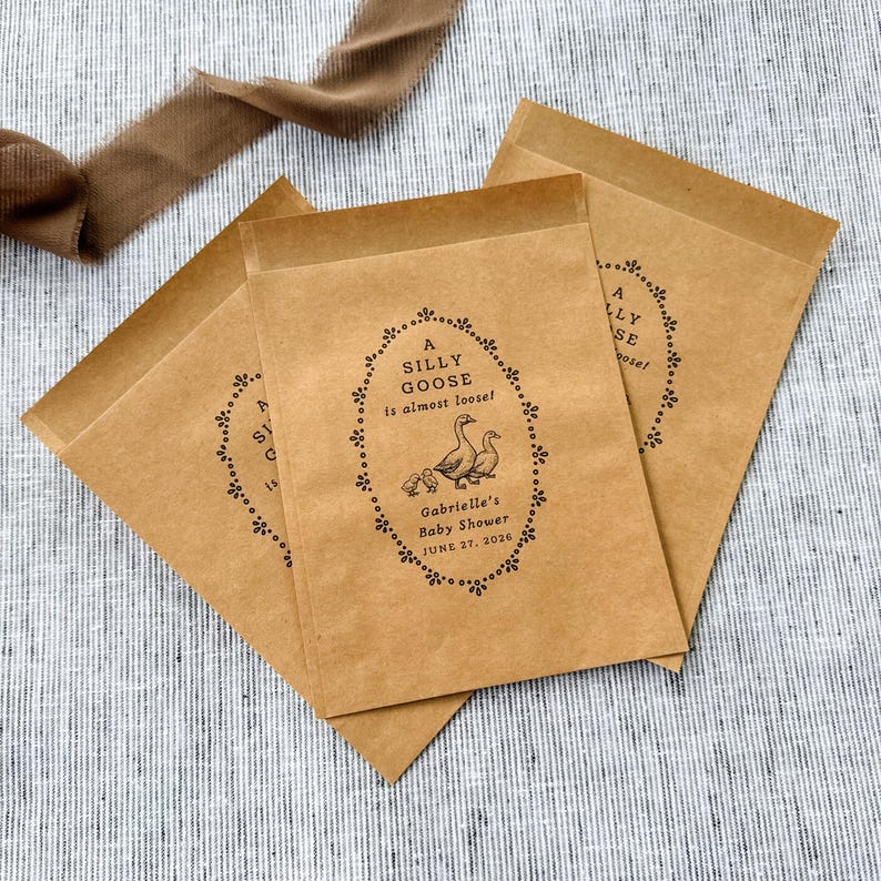 Puede incluir: Tres bolsas de papel marr&oacute;n para regalos con el texto "A SILLY GOOSE is almost loose!" en un marco ovalado decorativo. El marco incluye ilustraciones de gansos y gansitos. El texto "Gabrielle's Baby Shower JUNE 27, 2026" tambi&eacute;n est&aacute; impreso en las bolsas.