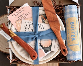 May include: A sourdough bread baking kit in a box containing a round banneton basket, a wooden lame with a metal blade, a wooden spreader, and a blue and white checkered towel labeled Salted Butter. Includes printed cards with text.