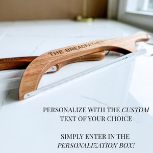 May include: A wooden bread slicer with a stainless steel blade. The words "THE BREADFATHER" are engraved on the wooden handle. The image also includes the text "PERSONALIZE WITH THE CUSTOM TEXT OF YOUR CHOICE" and "SIMPLY ENTER IN THE PERSONALIZATION BOX!"
