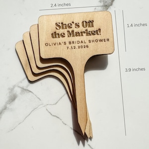 May include: A stack of four wooden plant markers, each measuring approximately 2.4 inches wide and 3.9 inches tall. The markers have a rectangular top with a handle. Text on the top reads "She's Off the Market!" and "OLIVIA'S BRIDAL SHOWER 7.12.2025".