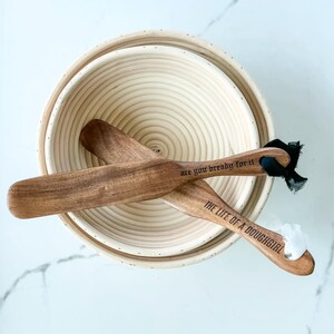 May include: Two wooden bread lame tools with the text "are you bready for it" and "THE LIFE OF A DOUGHGIRL" are crossed over a round, woven proofing basket. The tools have black ribbon and white material attached.