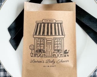 以下が含まれることがあります： CAFÉ BÉBÉと書かれた店舗の黒いインクのイラストが描かれた茶色の紙袋。テーブル、椅子、ベビーカーが描かれています。店舗の下にはLauren's Baby Showerと日付18.10.2027の文字があります。上部には黒いサテンのリボンが結ばれています。