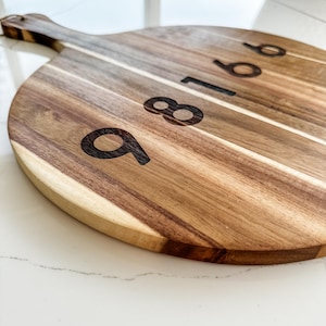 May include: A round wooden chopping board with a handle. The board has the numbers 9, 8, 1, 6, and 8 engraved on it.
