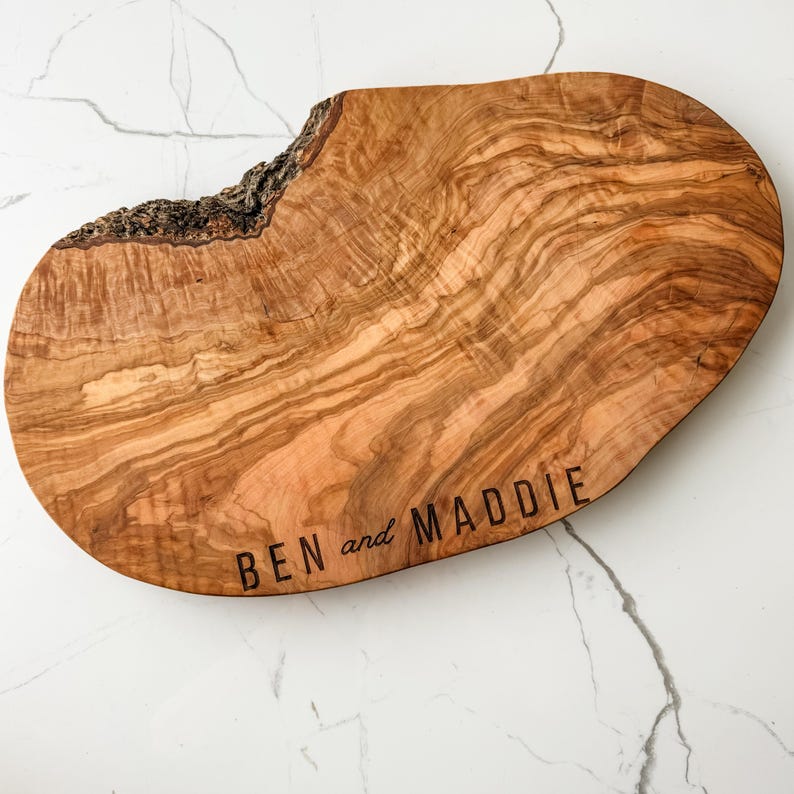 May include: A wooden serving board with a natural edge and a light brown finish. The board has the engraved text "BEN and MADDIE" in a simple font. The board is set against a white marble background.