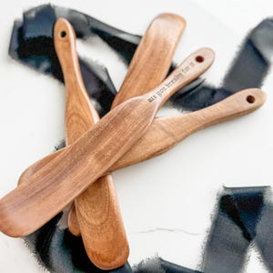 May include: Several wooden bread spreaders with a natural finish are arranged on a white surface. One spreader has the phrase "are you bready for it" etched into the handle. Each spreader has a hole at the end of the handle.