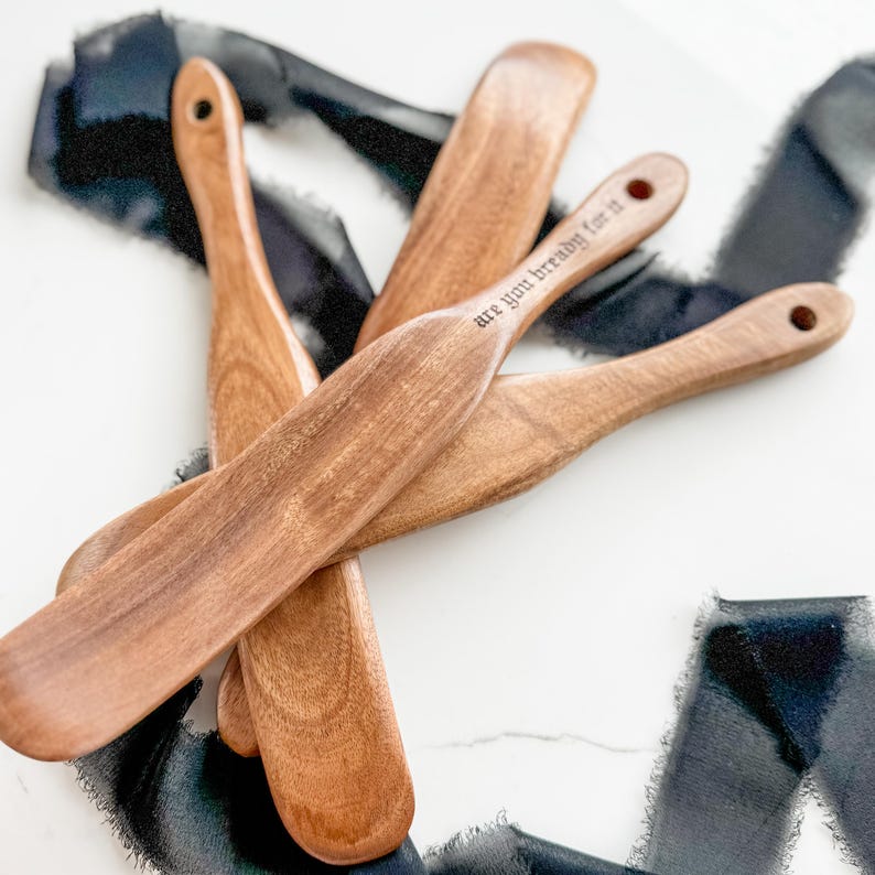 May include: Several wooden bread spreaders with a natural finish are arranged on a white surface. One spreader has the phrase "are you bready for it" etched into the handle. Each spreader has a hole at the end of the handle.