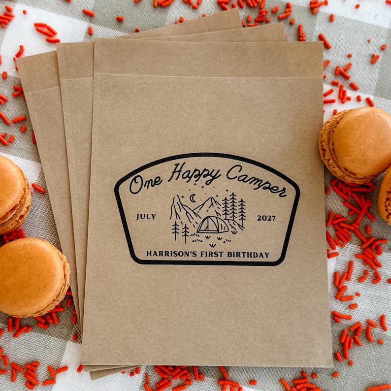 May include: A stack of light brown paper bags featuring a black printed design with a mountain, tent, and pine tree graphic. The text reads One Happy Camper, July 2027, and Harrison's First Birthday, arranged in a badge-like shape on the front.