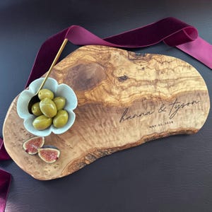 May include: A wooden serving board with a natural edge, engraved with "hanna & tyson" and the date May 22, 2026. It holds a small, flower-shaped bowl with green olives, a gold spoon, and fig slices. A burgundy ribbon is draped across the board.
