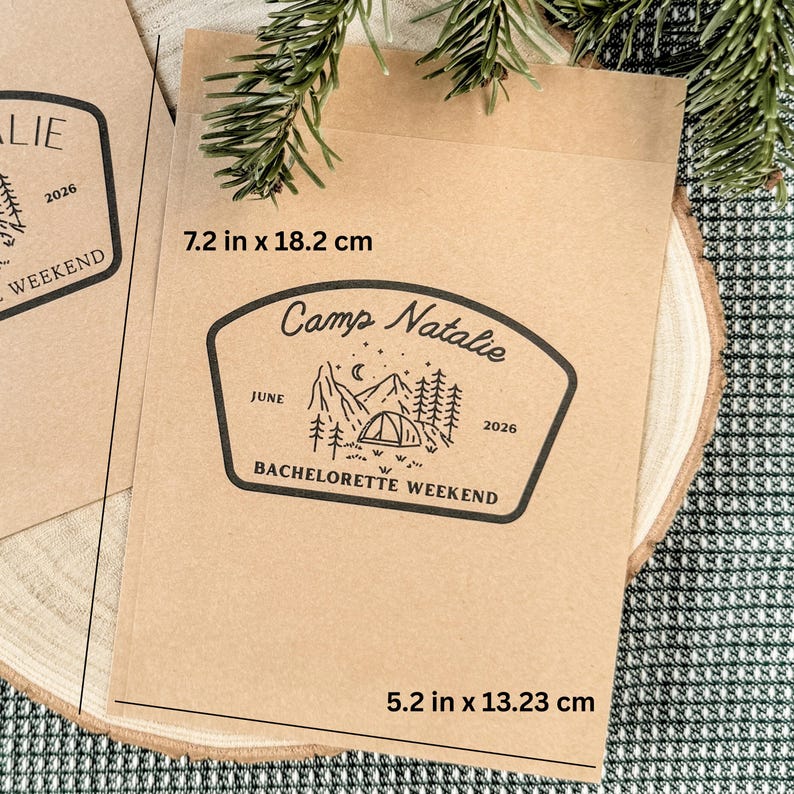 Pode incluir: Um saco de papel castanho retangular com um gr&aacute;fico impresso a preto de uma paisagem montanhosa, uma tenda e pinheiros. O texto diz Camp Natalie, June 2026 e BACHELORETTE WEEKEND. As dimens&otilde;es s&atilde;o 7,2 polegadas por 18,2 cm e 5,2 polegadas por 13,23 cm.