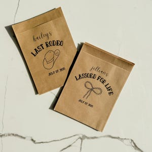 May include: Two brown paper favor bags with black text and illustrations. One bag reads "hailey's LAST RODEO" with a cowboy hat graphic and the date "JULY 27, 2025." The other bag says "jillians LASSOED FOR LIFE" with a lasso graphic and the same date.