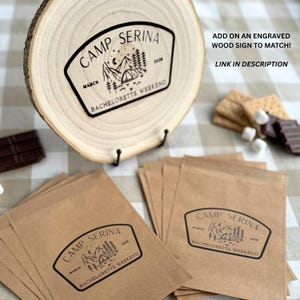 May include: A round wood slice sign and several brown paper bags, each featuring a black printed graphic of a tent, mountains, trees, and the text CAMP SERINA MARCH 2026 BACHELORETTE WEEKEND. The image also includes text about an engraved wood sign.