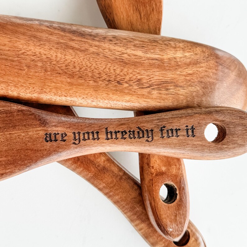 May include: Wooden kitchen utensils, including a spatula and a spoon, are displayed. The spatula has the phrase "are you bready for it" etched into the wood. The utensils are a warm brown color.