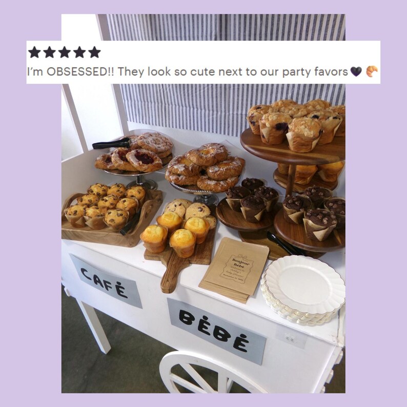 May include: A white cart labeled CAF&Eacute; B&Eacute;B&Eacute; displays an assortment of baked goods including muffins, pastries, and cookies on wooden trays and tiered stands. A stack of white plates and small paper bags with the text Bonjour b&eacute;b&eacute; are also present.