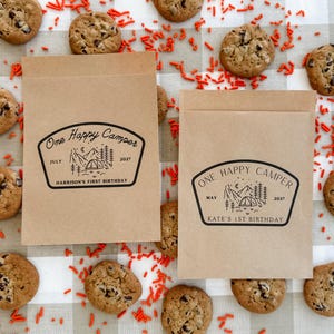 May include: Two rectangular brown paper bags featuring a black ink stamp design of a mountain landscape with a tent, trees, and moon. The text reads One Happy Camper, July 2027, Harrison's First Birthday and One Happy Camper, May 2027, Kate's 1st Birthday.