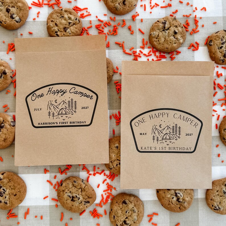 May include: Two rectangular brown paper bags featuring a black ink stamp design of a mountain landscape with a tent, trees, and moon. The text reads One Happy Camper, July 2027, Harrison's First Birthday and One Happy Camper, May 2027, Kate's 1st Birthday.