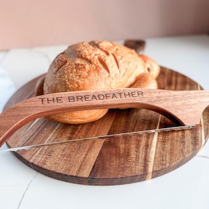 May include: A crusty loaf of bread rests on a wooden bread board, with a wooden bread slicer in place. The slicer is engraved with the words "THE BREADFATHER". The round bread board is made of wood and has a diameter of approximately 30 cm.