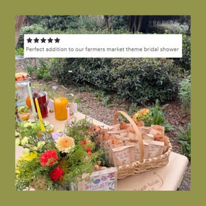May include: A woven wicker basket filled with small paper bags containing pastries, each labeled with the handwritten text 'Fresh Off The Market!'. A colorful floral arrangement, glass pitchers of orange and red juice, and candles sit nearby.