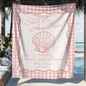 May include: A rectangular fabric banner featuring a light pink and white gingham border. The centre displays a large pink seashell illustration with the text "She'll Be Here Soon!", "Welcome to Cindy and Daryn's Baby Shower", and "August 15th, 2026".