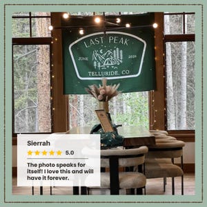 May include: A rectangular dark green wall banner featuring white line art of mountains, pine trees, a tent, and the text LAST PEAK, JUNE 2026, TELLURIDE, CO. The banner is displayed indoors above a wooden table with chairs and a floral arrangement.