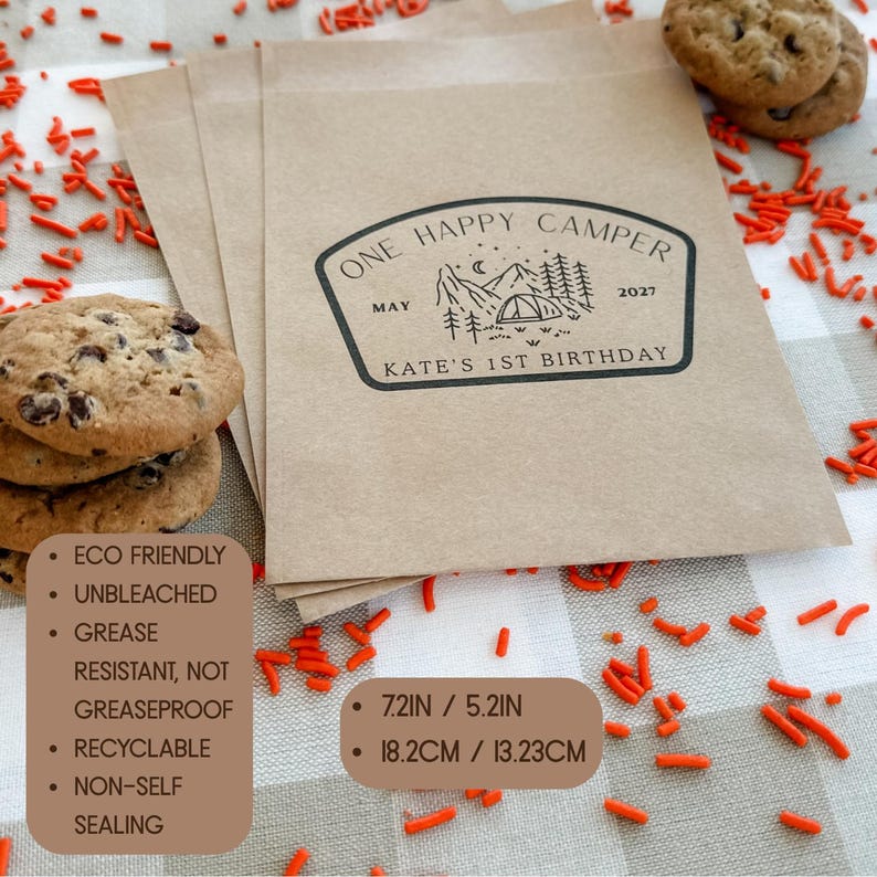 May include: A stack of light brown paper bags featuring a black ink stamp with a mountain, tent, and tree graphic. The text reads ONE HAPPY CAMPER, MAY 2027, and KATE'S 1ST BIRTHDAY. The bags measure 7.2 inches by 5.2 inches and are unbleached.