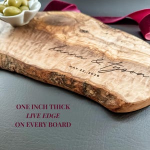 May include: A wooden serving board with a live edge, engraved with the names "hanna & Tyson" and the date "MAY 22, 2026". The board is approximately one inch thick. A small white bowl of green olives sits on the top left. A burgundy ribbon is in the background.