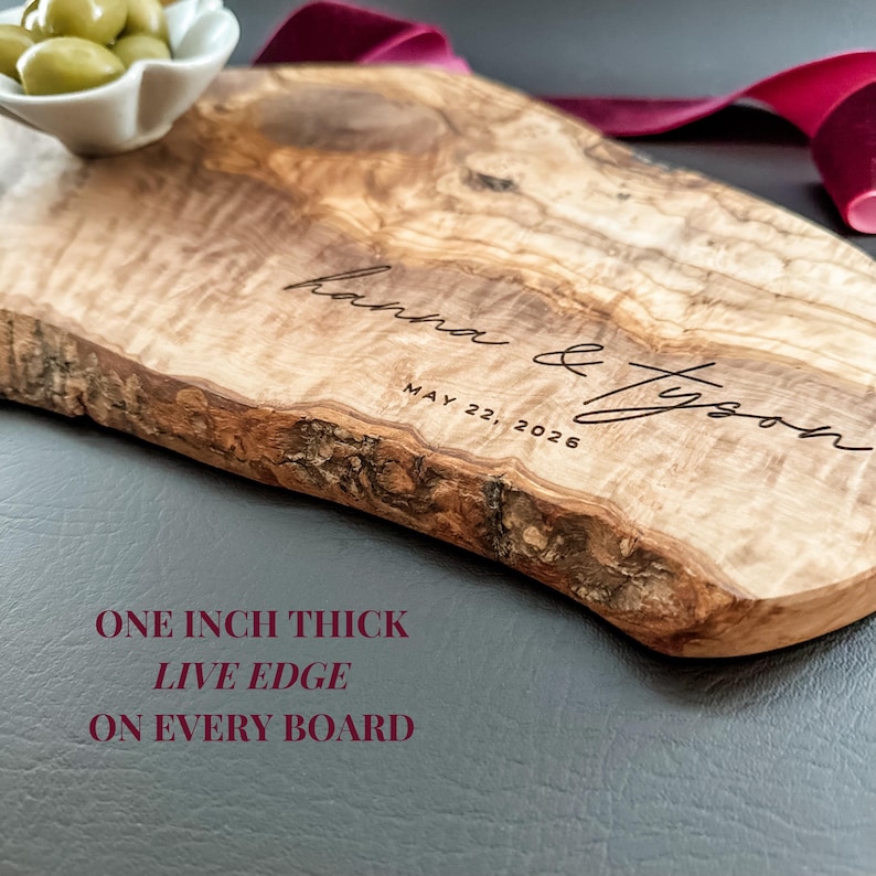May include: A wooden serving board with a live edge, engraved with the names "hanna & Tyson" and the date "MAY 22, 2026". The board is approximately one inch thick. A small white bowl of green olives sits on the top left. A burgundy ribbon is in the background.