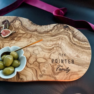 May include: A wooden serving board with a natural wood grain pattern. The board features the text "The Pointer Family". A small bowl of olives, figs, and a gold spoon are on the board. A burgundy ribbon is in the background.