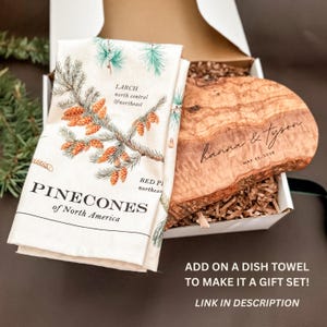 May include: A gift set featuring a white dish towel with a pinecone design and a wooden cutting board. The towel has text that reads "PINECONES of North America". The cutting board is engraved with a name and date. The set is presented in a white box.