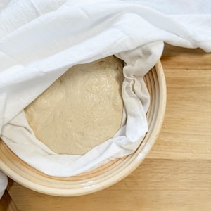 Puede incluir: Un pa&ntilde;o blanco cubre una bola de masa redonda y de color marr&oacute;n claro en una cesta tejida. La cesta est&aacute; sobre una superficie de madera.