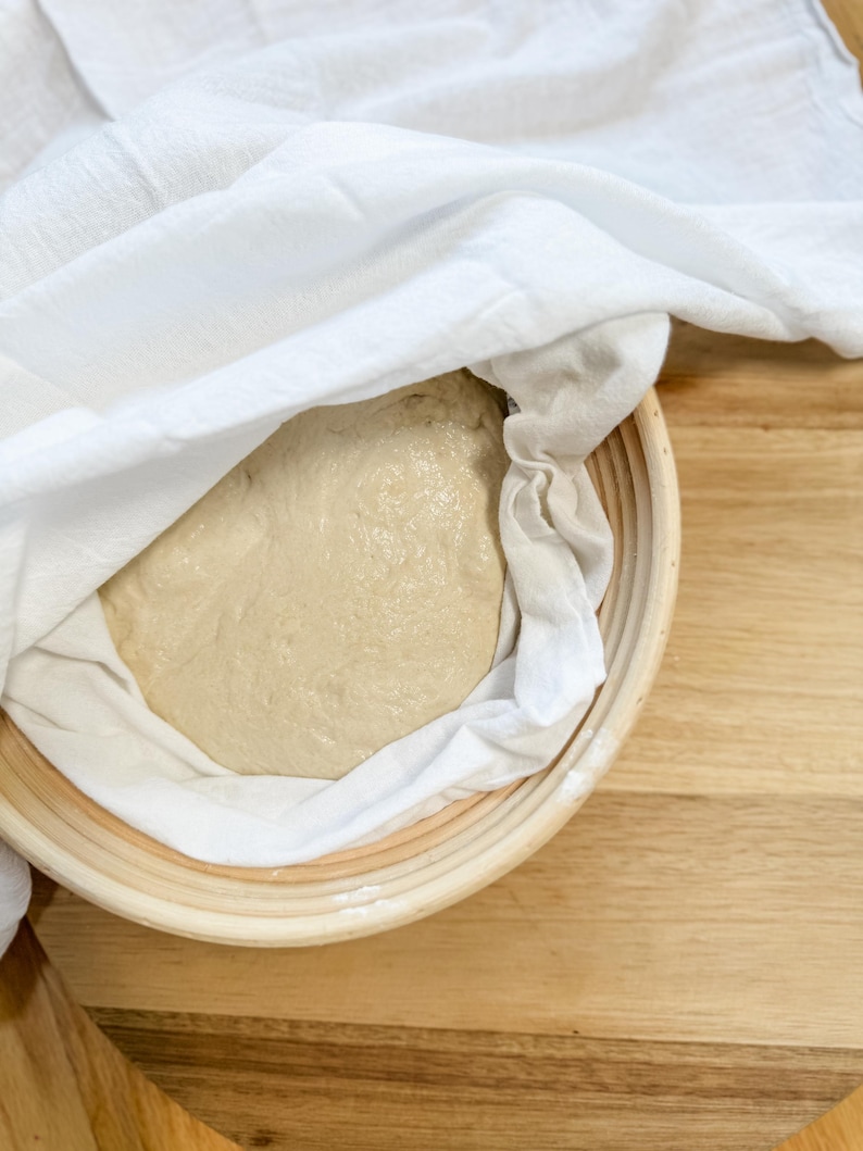 Puede incluir: Un pa&ntilde;o blanco cubre una bola de masa redonda y de color marr&oacute;n claro en una cesta tejida. La cesta est&aacute; sobre una superficie de madera.