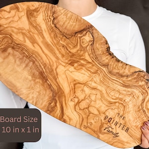 May include: A large, wooden cutting board with a natural wood grain pattern. The board is approximately 20 inches long, 10 inches wide, and 1 inch thick. The text "THE POINTER Family" is engraved on the board. The board is held by a person.