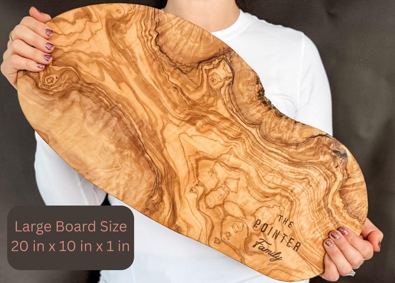 May include: A large, wooden cutting board with a natural wood grain pattern. The board is approximately 20 inches long, 10 inches wide, and 1 inch thick. The text "THE POINTER Family" is engraved on the board. The board is held by a person.