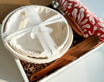 May include: A bread-baking kit in a white box. The kit includes two round, cream-colored proofing baskets tied with a white ribbon, a wooden spatula, a red and white patterned cloth, and a bread lame. The box is filled with brown paper.
