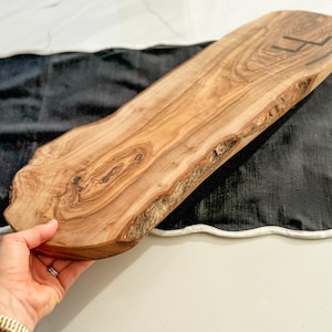 May include: A natural wood serving board with a live edge. The board is light brown with dark wood grain patterns and a dark initial on the top right. The board is held by a person's hand.