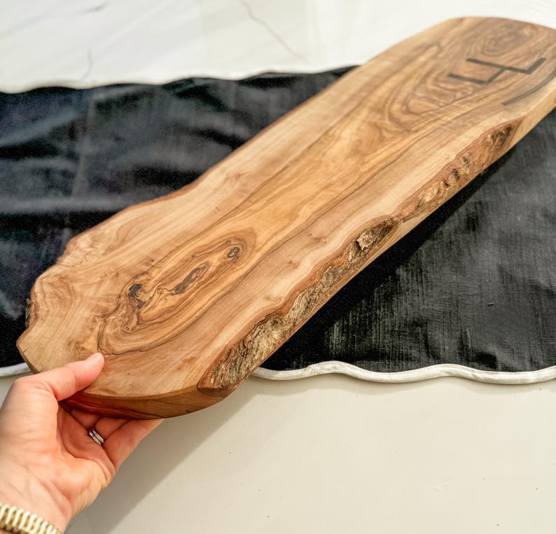 May include: A natural wood serving board with a live edge. The board is light brown with dark wood grain patterns and a dark initial on the top right. The board is held by a person's hand.
