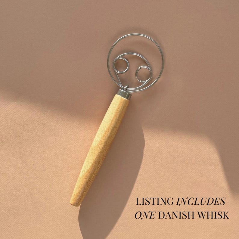 May include: A Danish whisk with a wooden handle and a silver metal wire loop. The whisk is on a light brown background. Listing includes one Danish whisk.