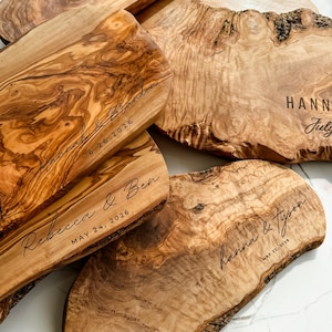 May include: A collection of wooden serving boards with natural edges. Each board is engraved with names and dates, such as "Hannah & Brandon 6.26.2026" and "Rebecca & Ben MAY 24, 2026". The boards have a warm, natural wood grain.
