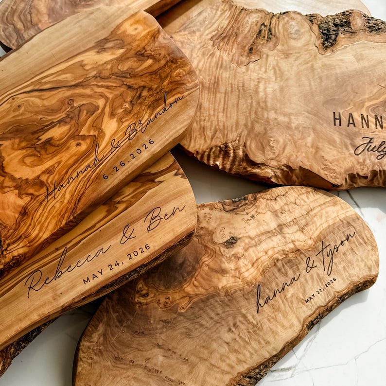 May include: A collection of wooden serving boards with natural edges. Each board is engraved with names and dates, such as "Hannah & Brandon 6.26.2026" and "Rebecca & Ben MAY 24, 2026". The boards have a warm, natural wood grain.