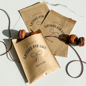 May include: Kraft paper bags with the text "jillian's LASSOED FOR LIFE" and the date "JULY 27, 2025" printed in black. The bags are filled with brown macarons. A brown leather cord is draped across the surface.