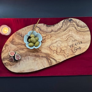 May include: An olive wood serving board with the text "The Pointer Family" engraved on it. The board holds a small blue bowl of olives, a gold spoon, and sliced figs. A lit candle sits to the left on a red surface.