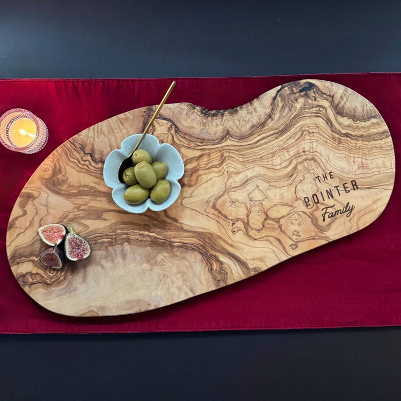 May include: An olive wood serving board with the text "The Pointer Family" engraved on it. The board holds a small blue bowl of olives, a gold spoon, and sliced figs. A lit candle sits to the left on a red surface.