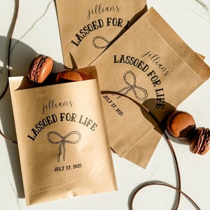 May include: Kraft paper bags with the text "jillians LASSOED FOR LIFE" and a rope design. The date "JULY 27, 2025" is printed on some bags. Several brown macarons are visible.