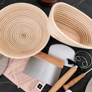 May include: A round and an oval rattan proofing basket, a metal dough scraper with a wooden handle, a white plastic bowl scraper, a metal dough whisk with a wooden handle, and a printed sourdough starter recipe card on a dark surface.