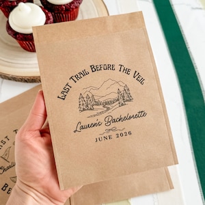 Op de afbeelding: Kraftpapieren cadeauzakjes met de tekst "Last Trail Before The Veil" en "Lauren's Bachelorette June 2026". De zakjes hebben een illustratie van een berg en bomen. De zakjes worden in een hand gehouden.