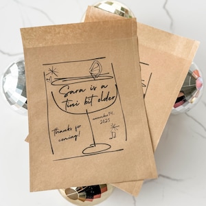 May include: Kraft paper favor bags with a black line drawing of a martini glass and the text "Sara is a tini bit older". The bags also include the date "november 14. 2025" and the phrase "thanks for coming!"