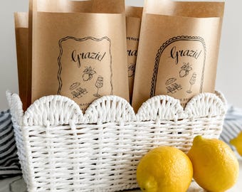 Può includere: Un cesto intrecciato bianco con bordo smerlato contiene diversi sacchetti di carta marrone stampati con la parola grazie, un'illustrazione di limone e icone da spiaggia. Due limoni giallo brillante sono posti davanti al cesto su una superficie bianca.