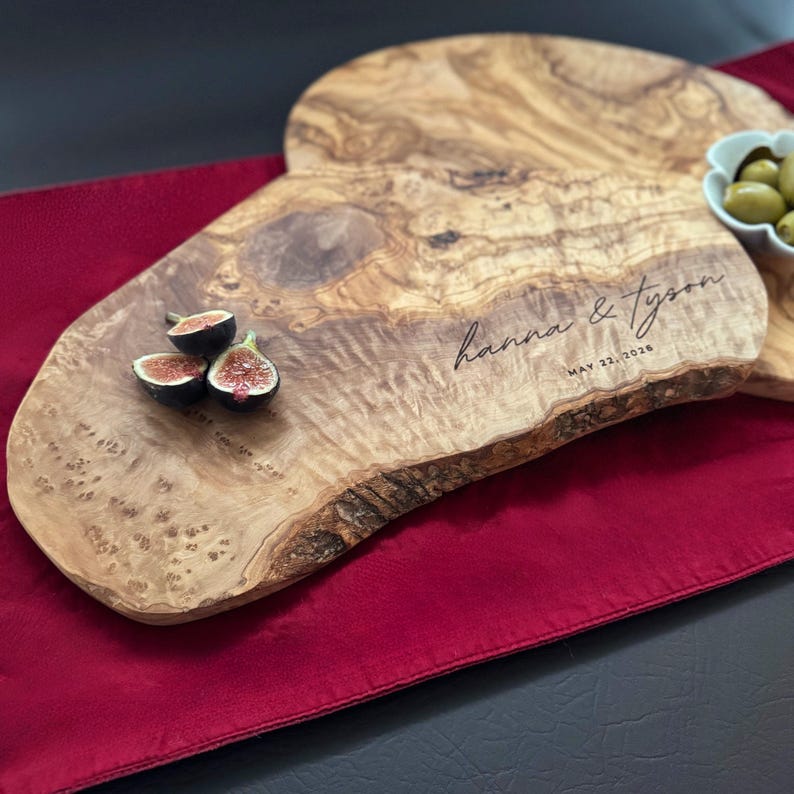 May include: Wooden serving boards with a natural edge, featuring the engraved names "hanna & tyson" and the date "MAY 22, 2026". The boards are adorned with figs and olives, set on a red tablecloth.